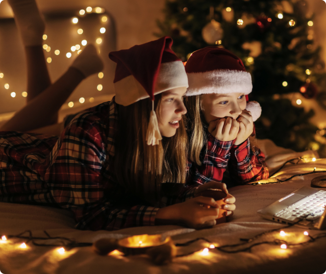 Zwei Mädchen folgen gespannt einem Weihnachtsfilm. Sie tragen gemütliche Pyjamas und Weihnachtsmützen.